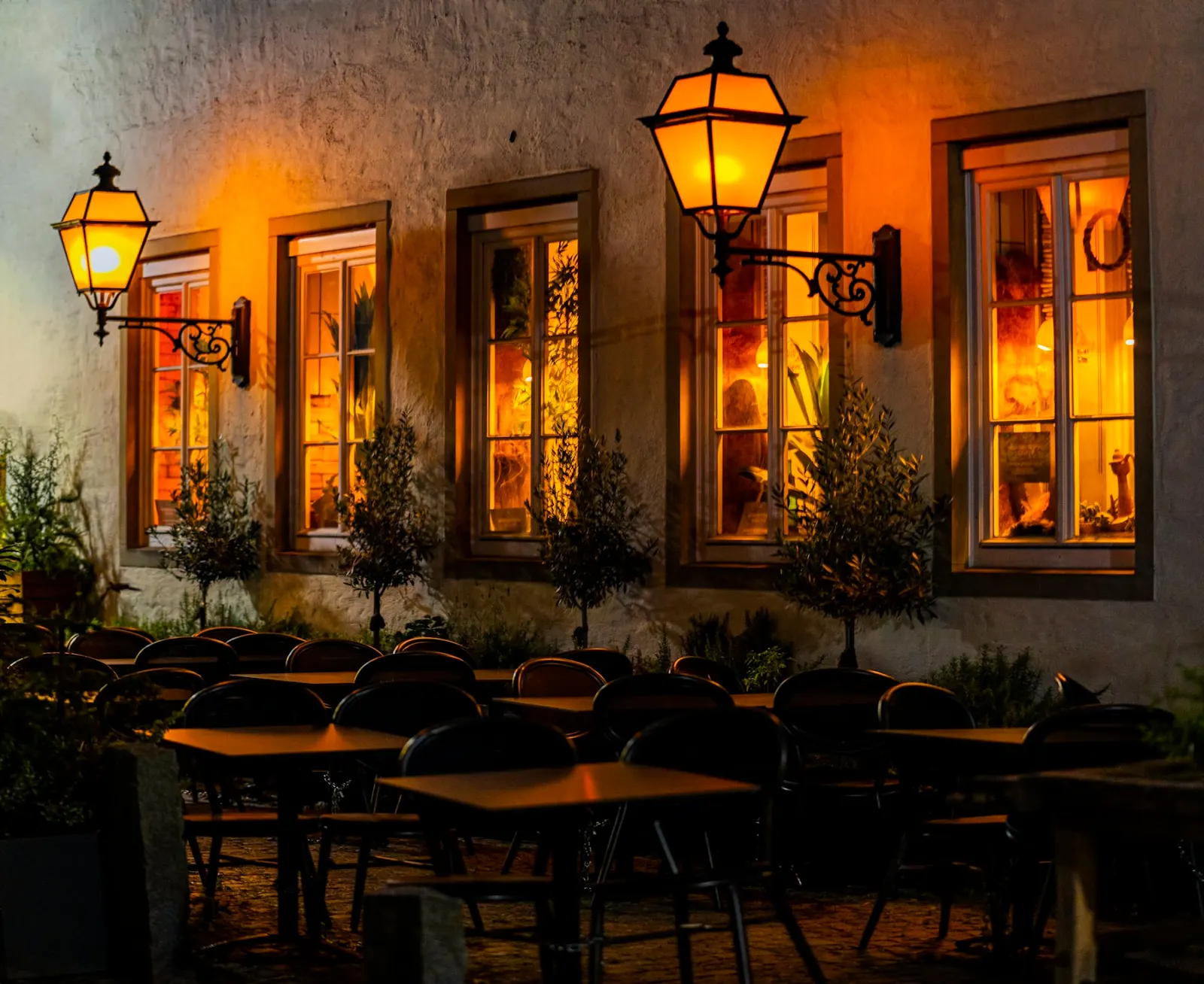 Večerní terasa trattorie s rozsvícenými lucernami a olivovníky pod okny